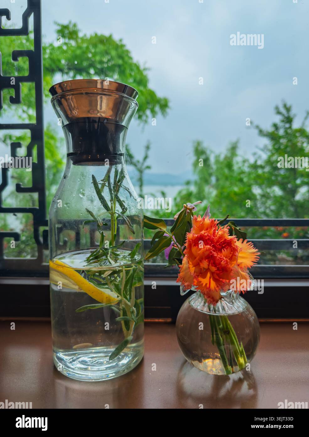 Caraffa limpida con acqua di limone rosmarino e fiori d'arancio che decorano un davanzale in legno che si affaccia su un tranquillo lago Foto Stock