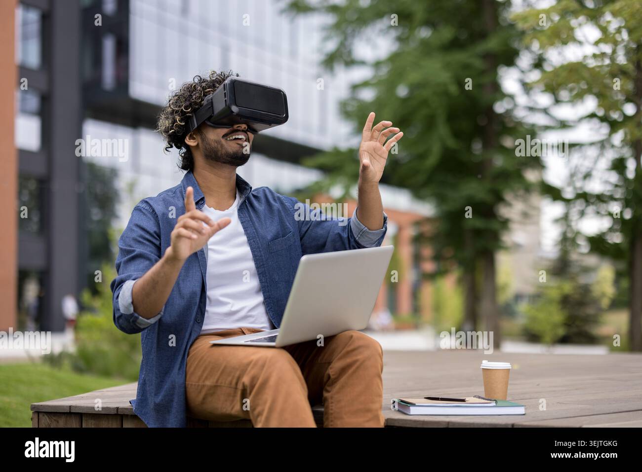 Giovane uomo che indossa un visore vr e tiene in mano un laptop, seduto all'aperto e interagisce con un ambiente virtuale utilizzando i gesti delle mani, sperimentando la realtà aumentata e l'innovazione digitale Foto Stock