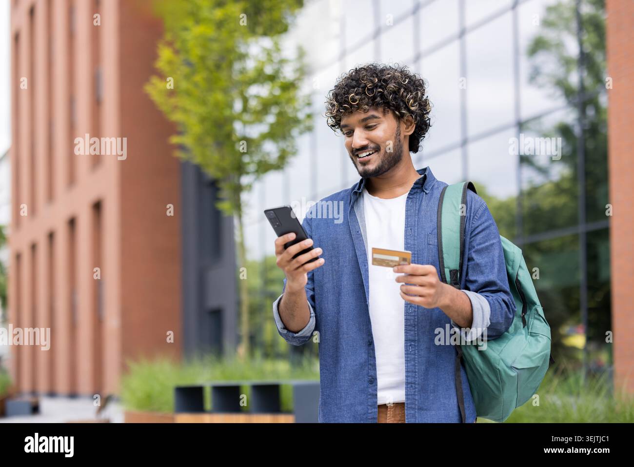 Giovane indiano con capelli ricci e zaino sorridente, che effettua un acquisto online utilizzando il suo smartphone e una carta di credito mentre si trova in un moderno campus urbano Foto Stock