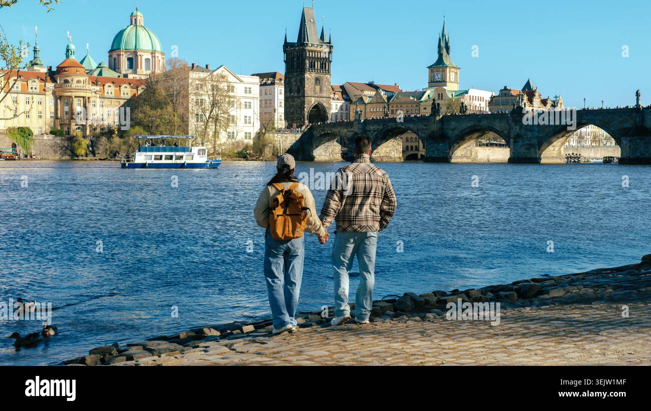 La coppia gode di una romantica passeggiata lungo il fiume Moldava, con splendide vedute dell'iconico skyline di Pragues. Il cielo azzurro e limpido esalta l'atmosfera vibrante, perfetta per una giornata memorabile di esplorazione. Foto Stock