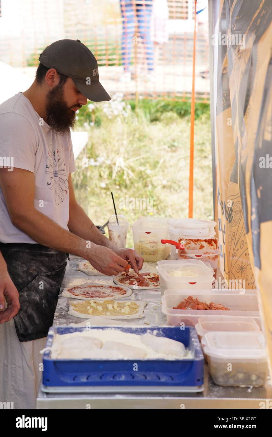 Venditore maschile barbuto che prepara pizza fresca al bancarello di cibo estivo all'aperto con vari condimenti e ingredienti nelle giornate di sole. Tonbridge Foto Stock