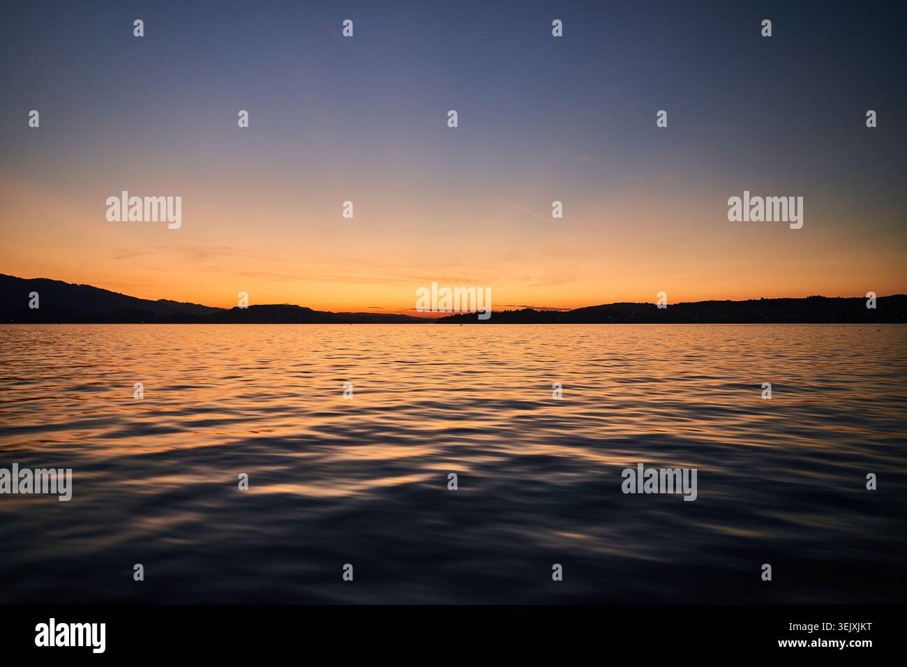 Il caldo tramonto dorato proietta riflessi su un lago tranquillo con colline sagomate e morbide increspature. Foto Stock