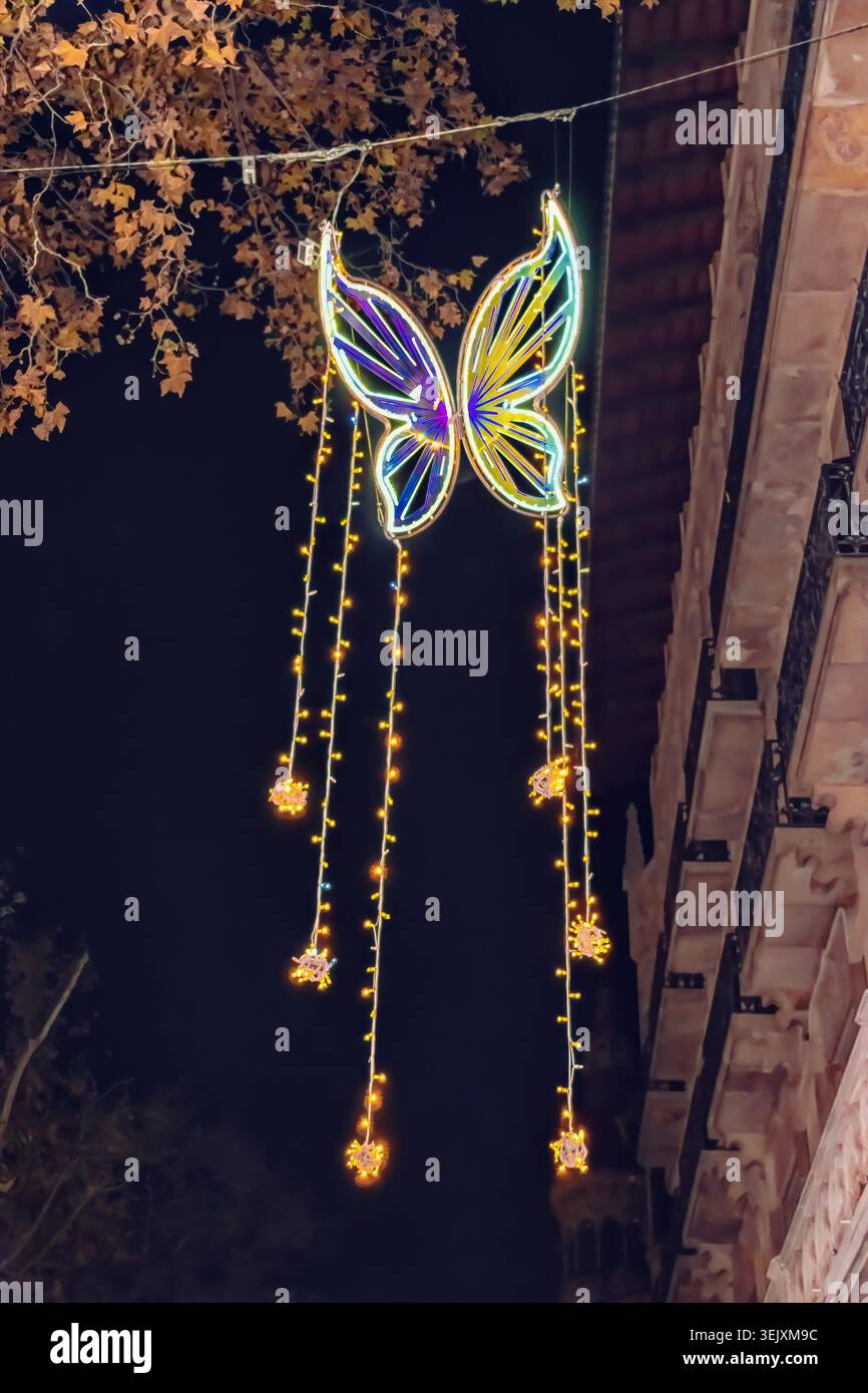 La farfalla blu al neon si illumina sullo sfondo di un albero con fogliame giallo e un cielo scuro al crepuscolo. Decorazioni natalizie sulla strada di Barcellona, Spagna Foto Stock