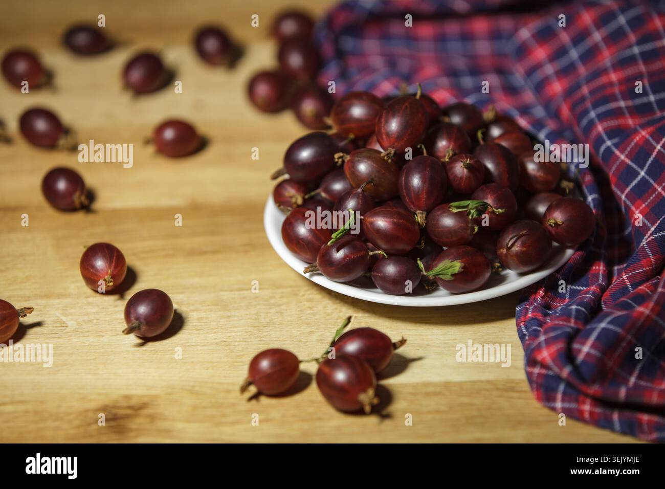 Piatto pieno di spina di colore rosso scuro maturo su un bordo di legno con frutti di bosco sparsi e un asciugamano da cucina a scacchi. Luce naturale calda, spirito di natura morta Foto Stock