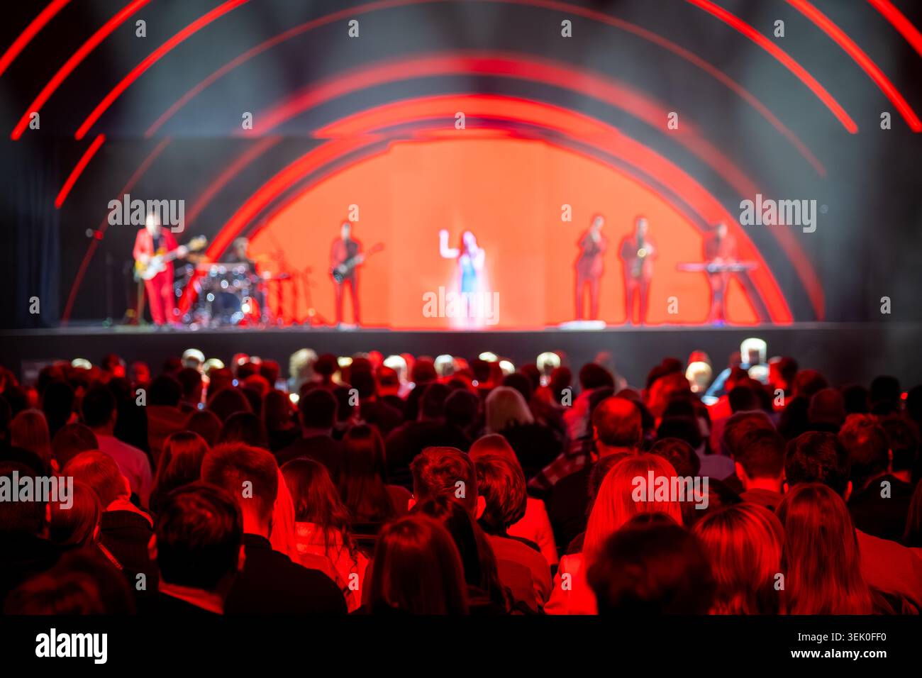Scena di concerti dal vivo con il pubblico di fronte al palco illuminato di rosso, band e cantante che si esibiscono, atmosfera energica. Foto Stock