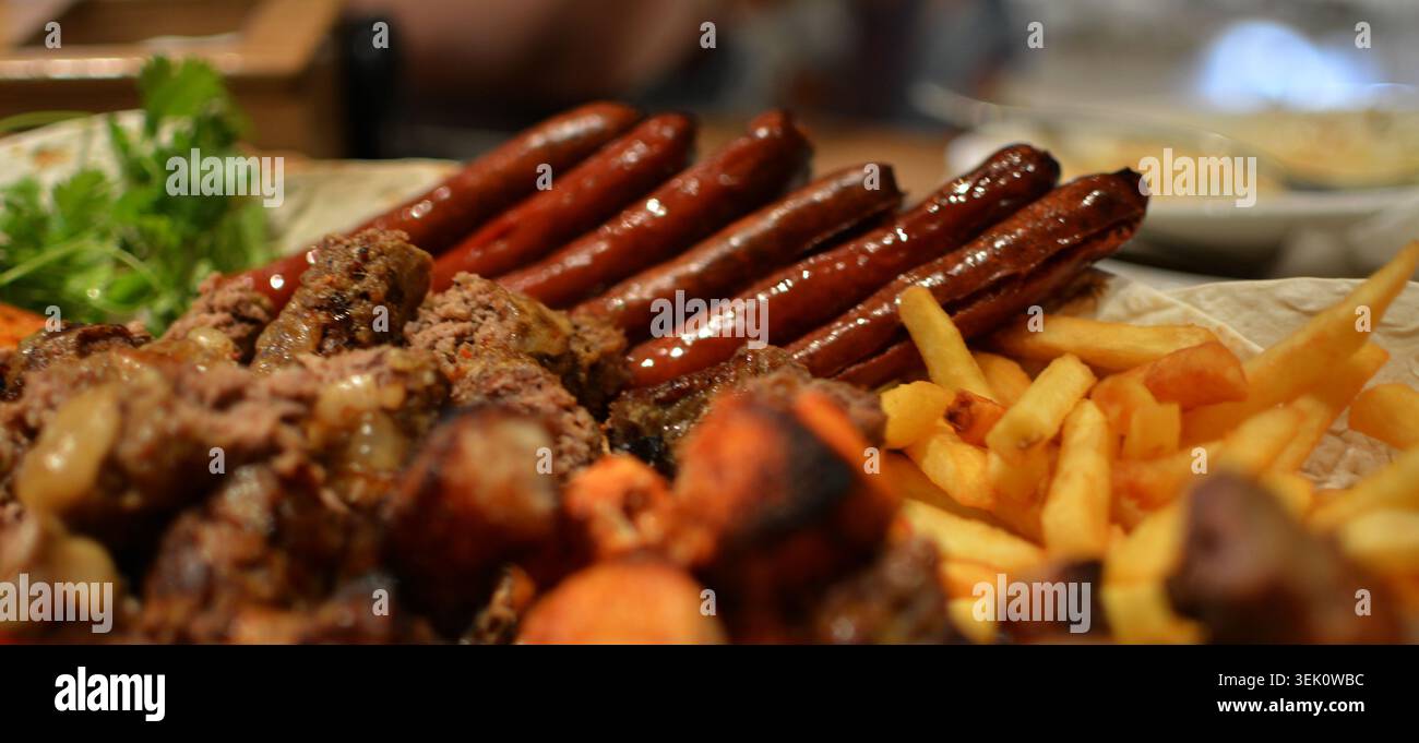 Primo piano di un abbondante piatto con salsicce grigliate, dettagli di carne, patatine fritte dorate croccanti e decorazioni fresche su un tavolo rustico con una calda luce soffusa Foto Stock