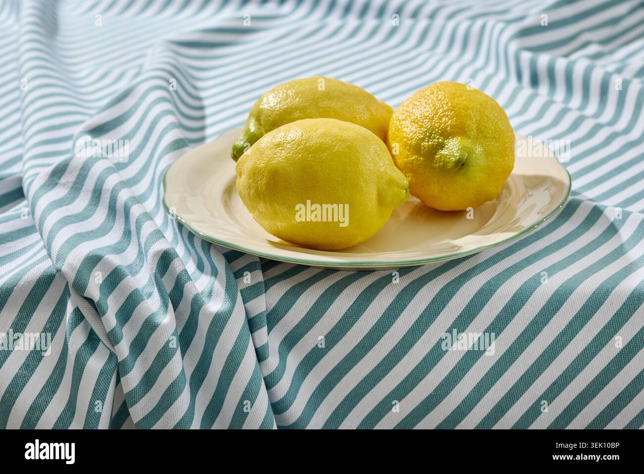 Foto a livello dell'occhio anteriore di tre limoni gialli vivaci collocati su un piatto in ceramica bianca dal design minimalista. La piastra si appoggia su un blu e un bianco ruvido e rugoso Foto Stock