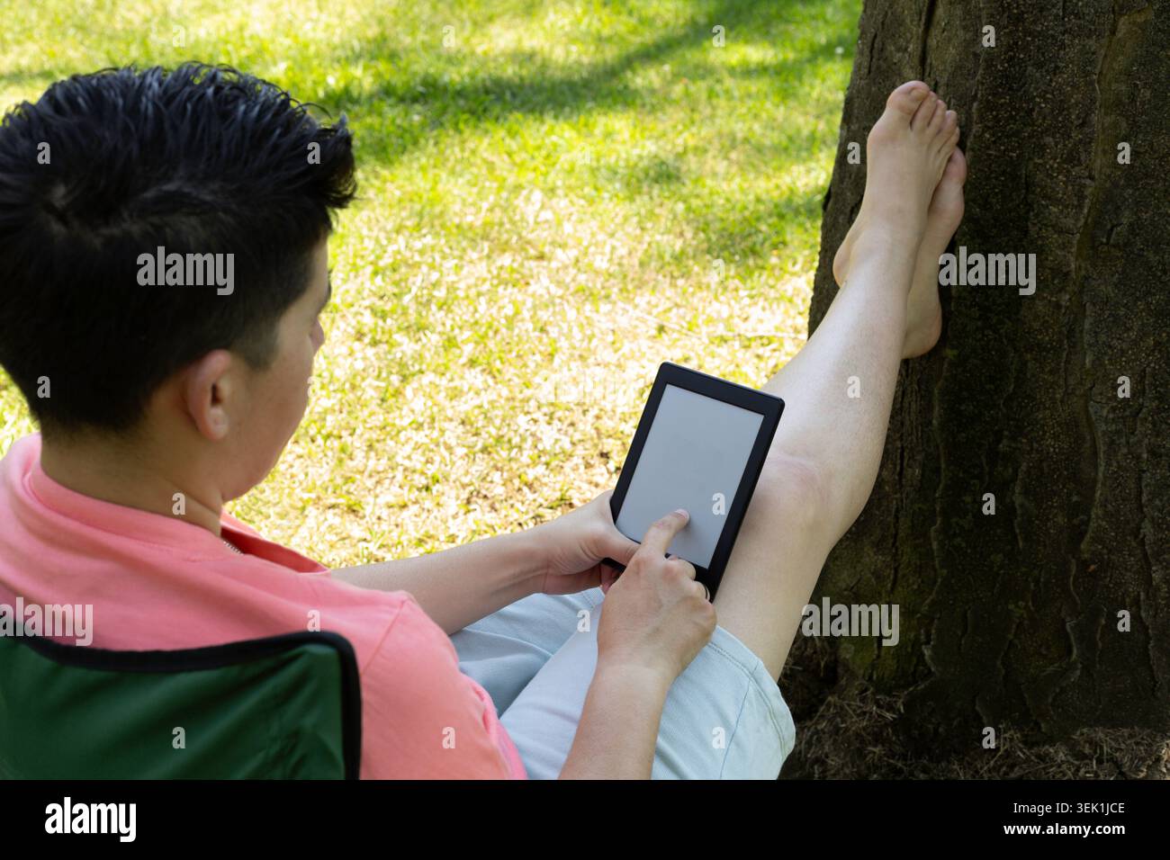 Un giovane uomo, con i piedi appoggiati contro un albero, sta leggendo un libro seduto su una sedia Foto Stock