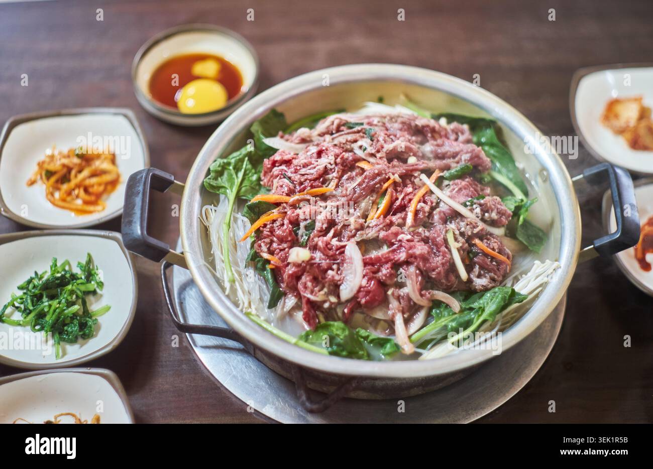 Bulgogi di manzo crudo fresco, spaghetti di vetro e verdure sono disposti in una pentola di metallo, accompagnati da tradizionali contorni coreani e una salsa di tuorli d'uovo. Foto Stock
