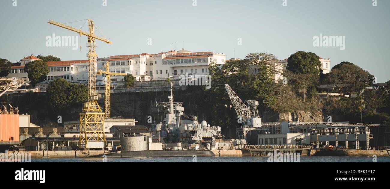 Sottomarino militare attraccato in un cantiere navale sotto le gru, con strutture industriali sul lungomare e un grande edificio bianco sulle colline nel pomeriggio Foto Stock