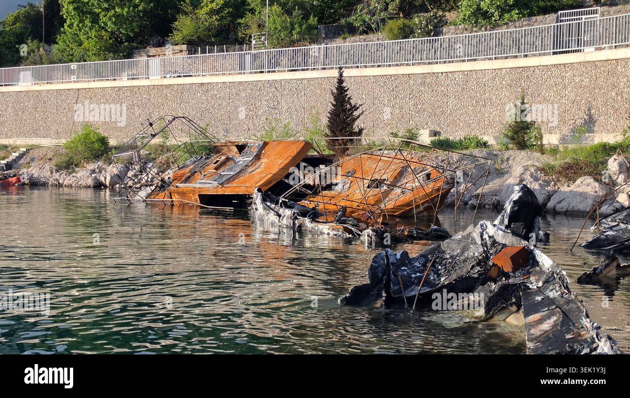 Un tiro medio cattura i resti arrugginiti e carbonizzati di una nave distrutta per metà sommersa in acque calme vicino a una costa rocciosa con una pietra recintata Foto Stock