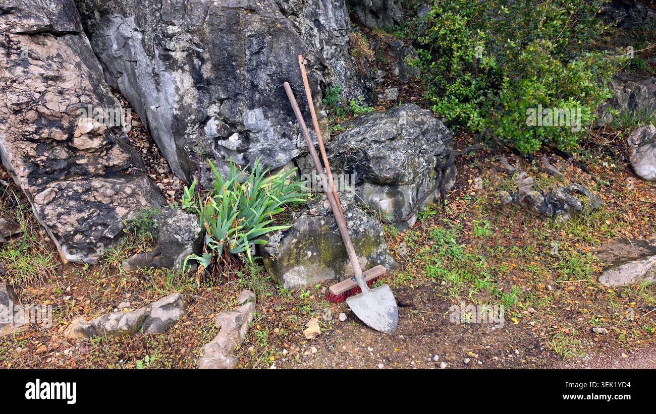 Una vanga di scavo in metallo intemprato e una lunga scopa a spinta in legno poggiano su massi calcarei naturali vicino a un lembo di Iris germanica barbuta verde Foto Stock