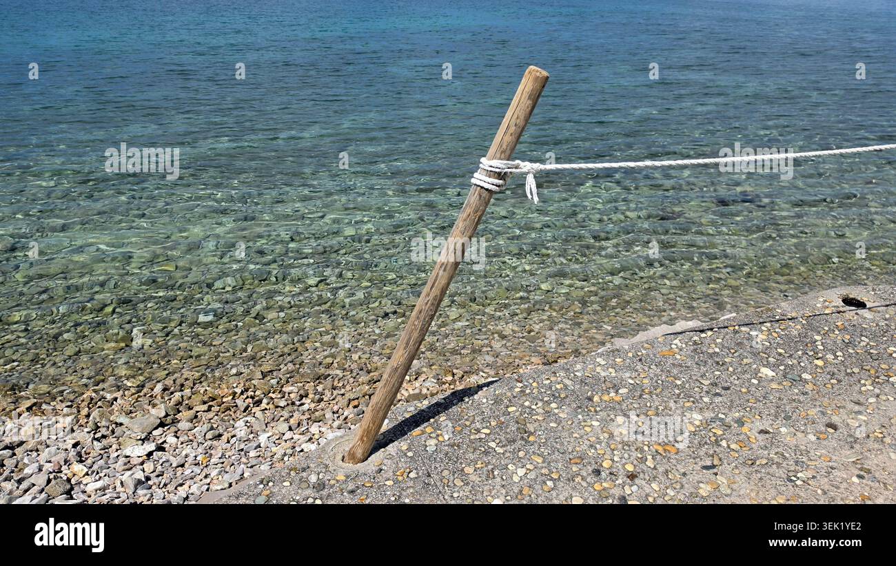 Un unico bastone di legno con una corda bianca avvolta intorno si erge su un bordo di cemento sopra una spiaggia di ciottoli limpidi Foto Stock