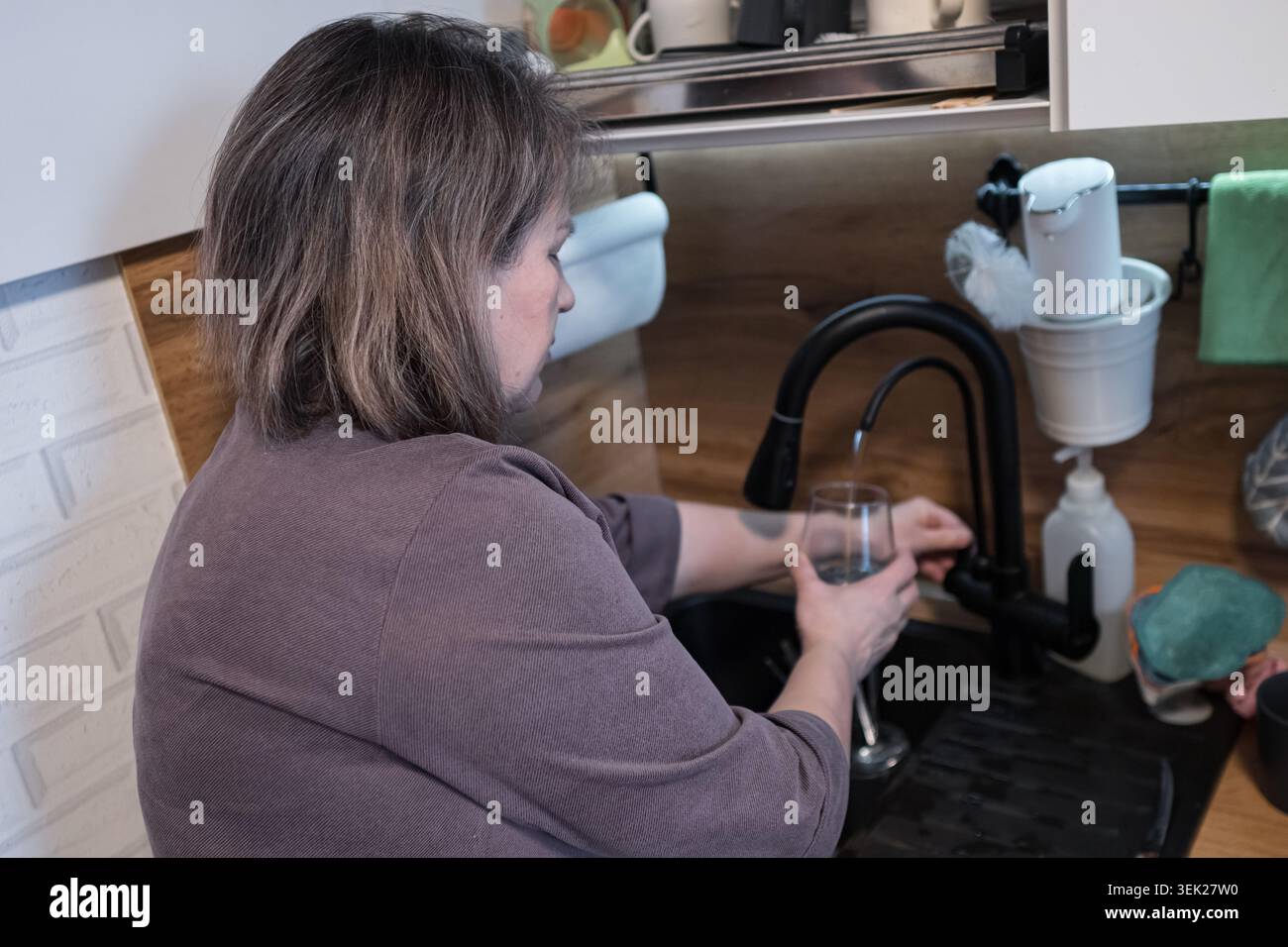Donna che lava un bicchiere in un lavandino in una casa moderna con scaffali aperti e articoli per la pulizia, rubinetto nero, spruzzi di legno, armadi bianchi Foto Stock
