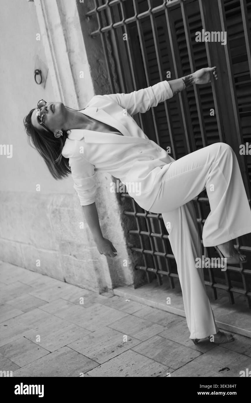 Donna con elegante abito bianco e occhiali da sole che si caratterizza per la sua dinamicità urbana poggia su una parete rustica di un edificio con grata in metallo, incarnando l'eleganza di un Foto Stock
