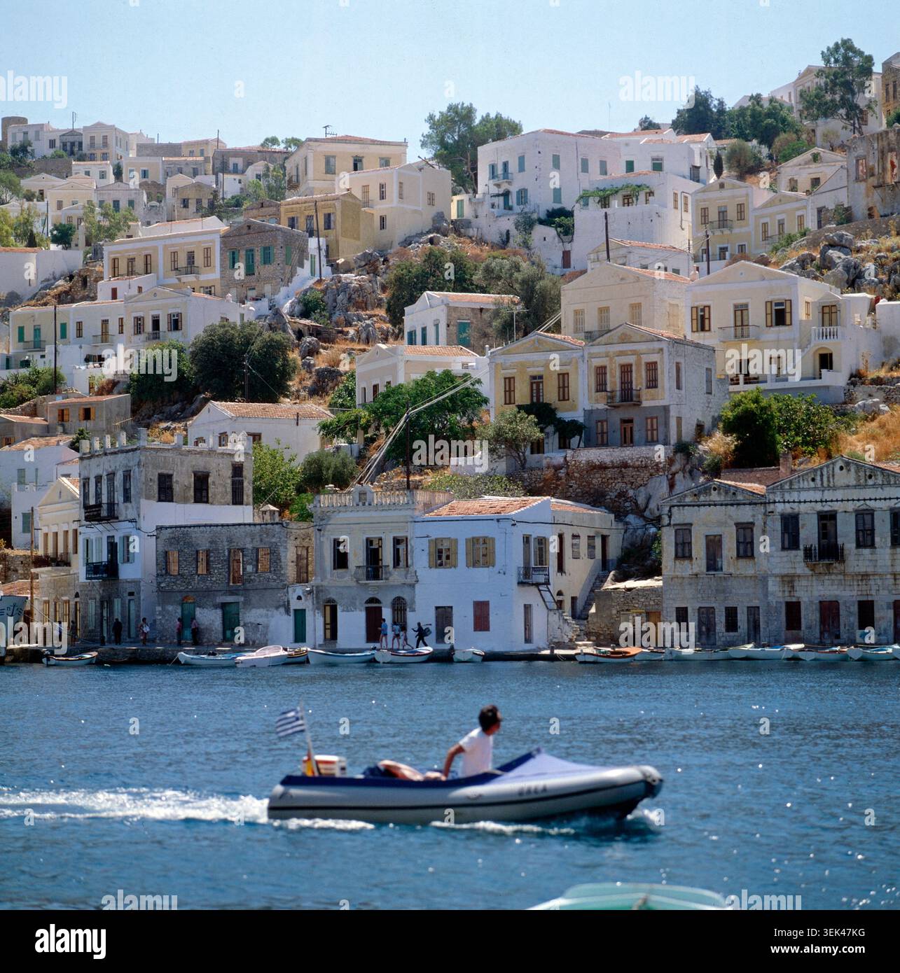 Ein malerischer Blick auf die Küstenstadt Symi in Griechenland, deren Häuser sich an einen Hügel schmiegen. IM Vordergrund fährt ein kleines Motorboot auf dem blauen Meer. Griechenland, um 1985. Foto Stock