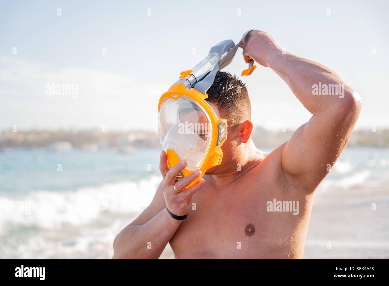L'uomo caucasico regola la maschera da snorkeling full face su una spiaggia con onde di mare e pelle bagnata durante le vacanze estive al sole. Snorkeling e attività ricreative marine. Foto Stock