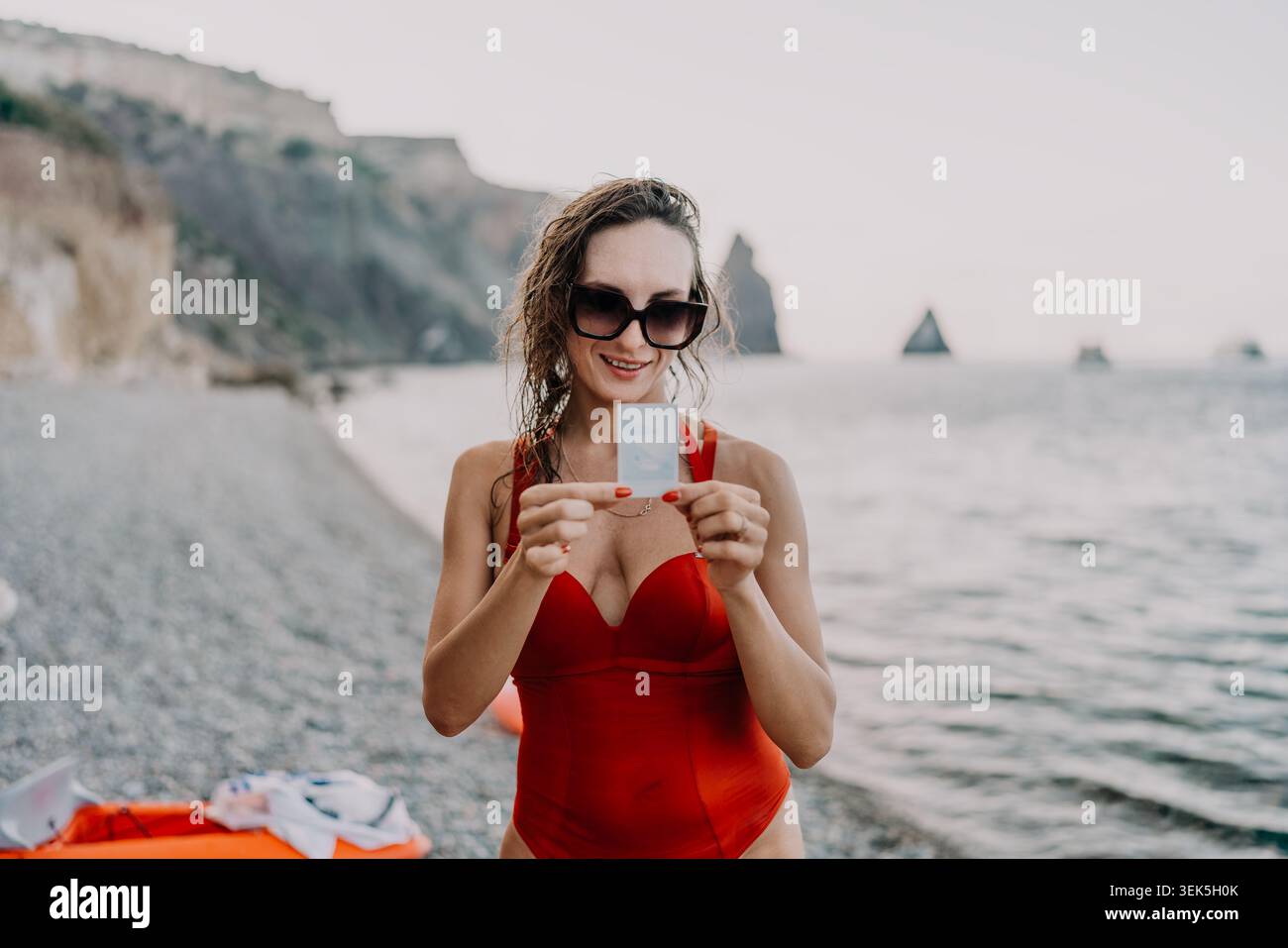 Donna, spiaggia, oceano. Donna sorridente in costume da bagno rosso con una piccola tessera su una tranquilla spiaggia di ciottoli. Foto Stock