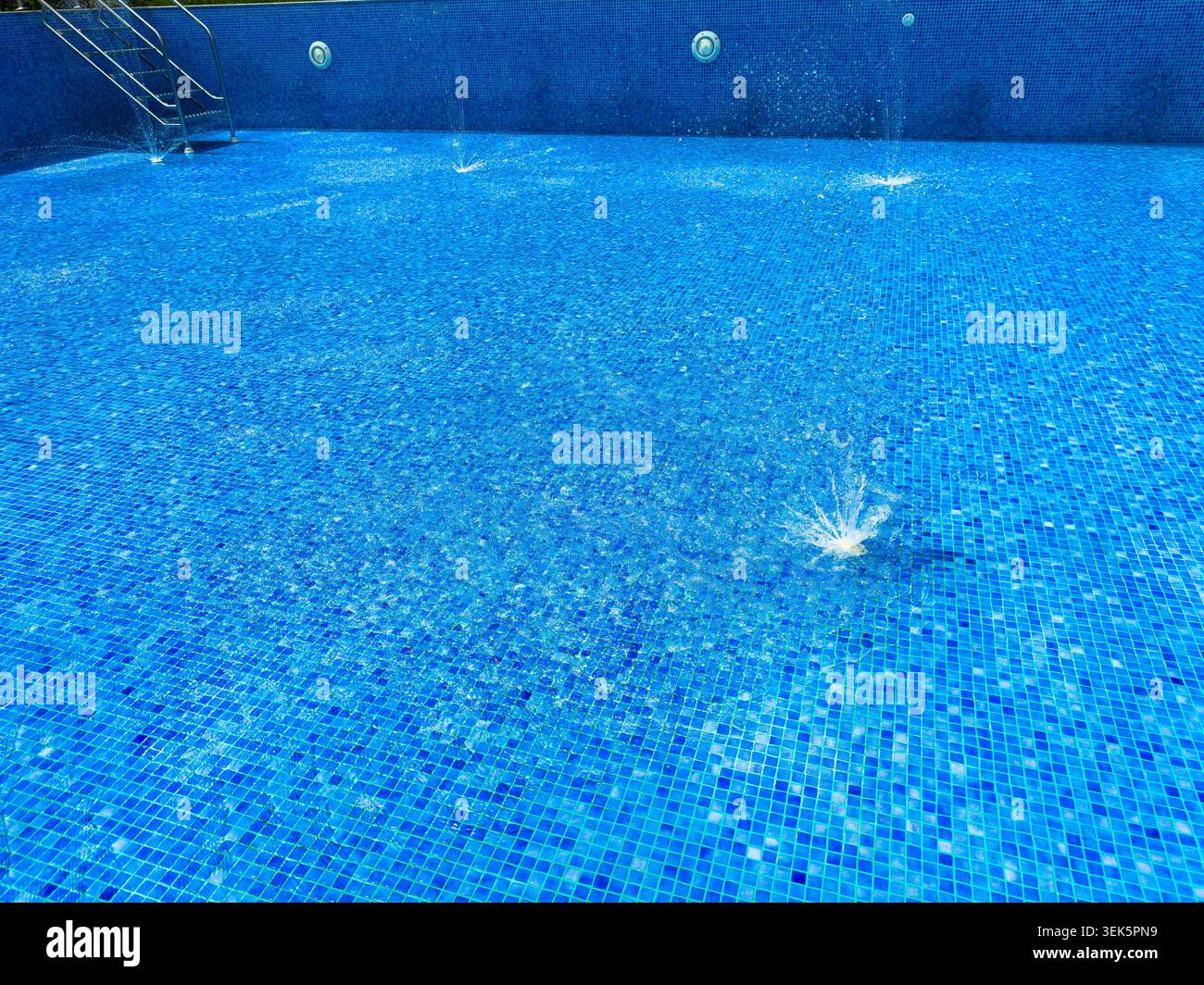 Gli spruzzi d'acqua provengono dal fondo della piscina. La luce del sole crea riflessi sulla superficie dell'acqua. Le piastrelle blu coprono l'area della piscina Foto Stock