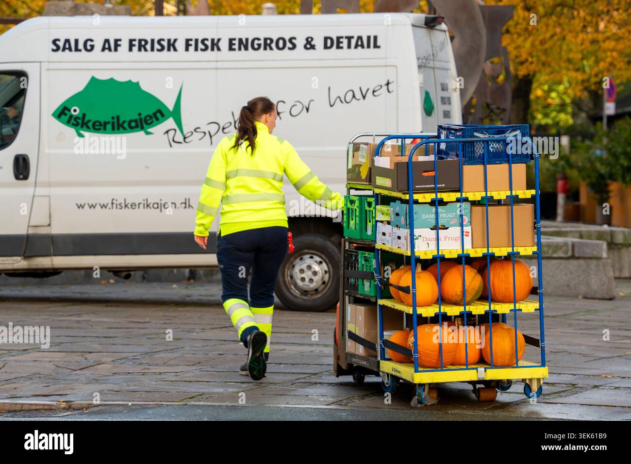 Una donna di colore giallo ad alta visibilità indossa un carrello elettrico per spostare due grandi carrelli di cibo, tra cui le grandi zucche arancioni di Halloween Foto Stock