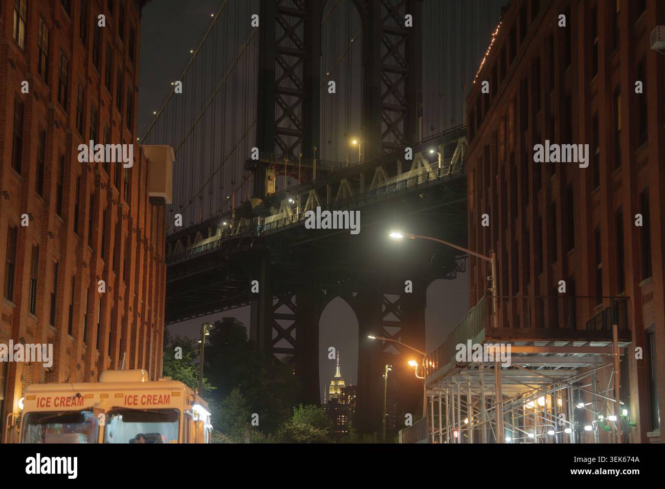 Strada notturna della città tra edifici in mattoni con un grande ponte sospeso, lampioni luminosi, ponteggi e un camion del gelato sotto. Foto Stock