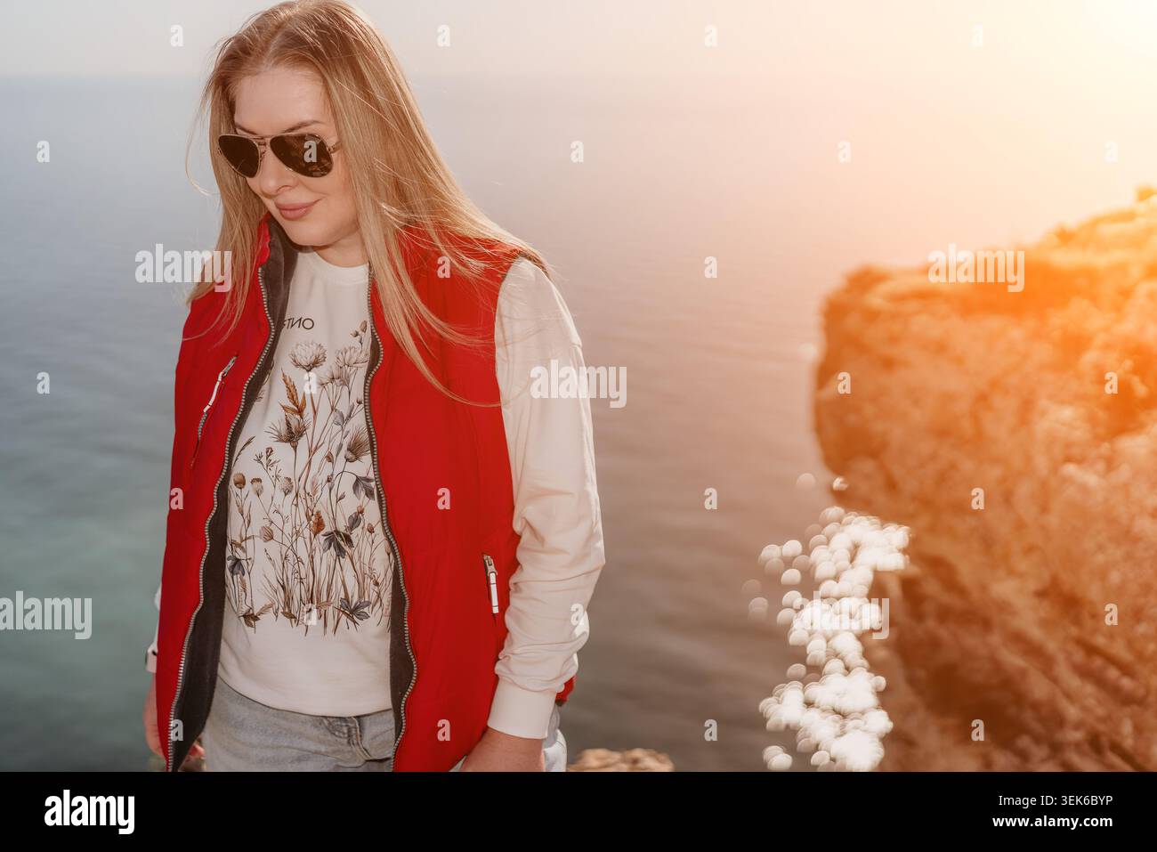 Donna bionda sulla scogliera del mare in gilet rosso in piedi sulla costa rocciosa che si affaccia sull'oceano all'ora d'oro con spazio fotocopie Foto Stock