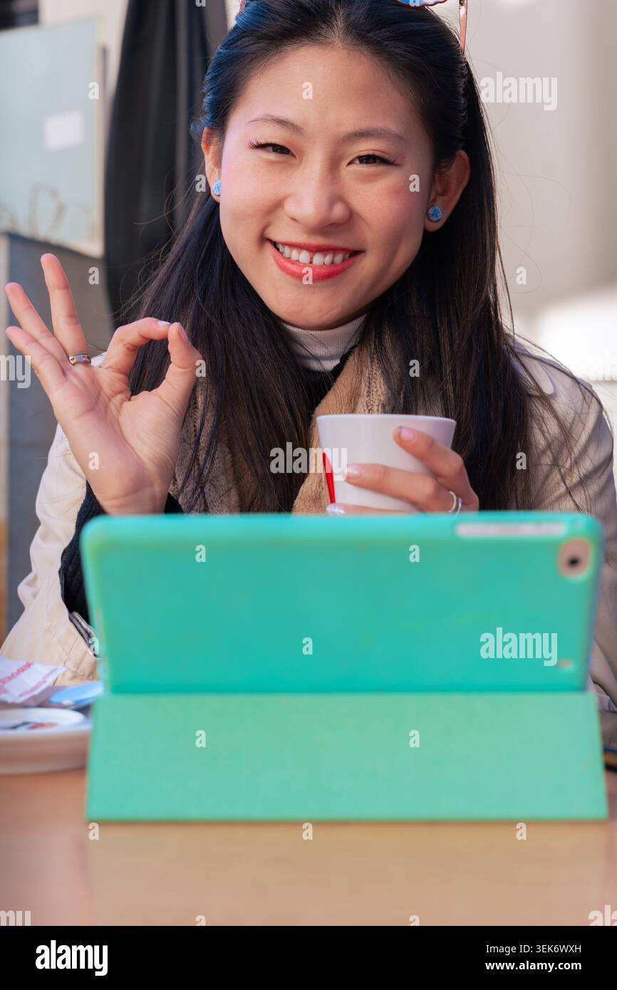 Donna sorridente che mostra un buon gesto tenendo il tablet e la tazza durante la pausa caffè. Verticale Foto Stock