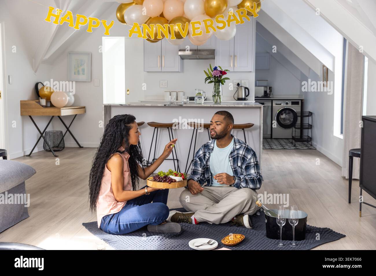 Coppie diverse sedute su una coperta scura nella cucina del loft, condividendo un vassoio di frutta in legno e champagne Foto Stock