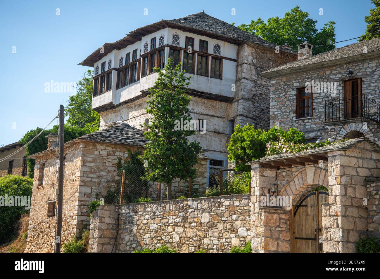 Una tradizionale casa di pietra illuminata dal sole con pareti bianche e accenti di legno scuro presenta finestre ornate e un tetto piastrellato contro un cielo blu luminoso. Foto Stock