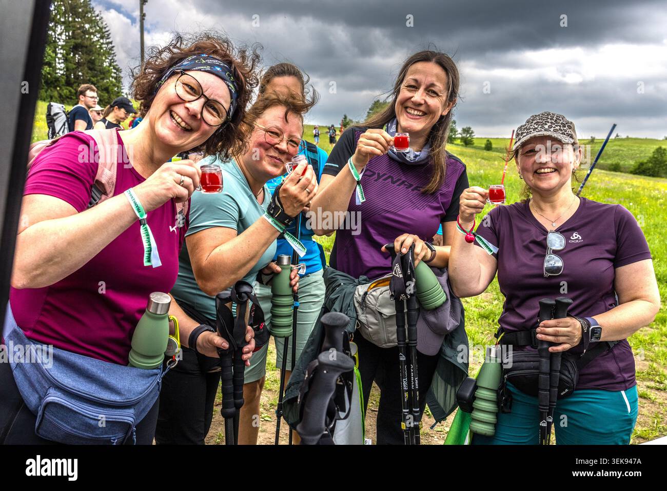 Quattro escursionisti sorridenti stringono bicchieri di schnapps in una sosta di ristoro durante la 14a maratona escursionistica di Frankenwald: Bastoni da passeggio in mano, un suggestivo cielo nuvoloso, Tettau, Baviera, Germania Foto Stock