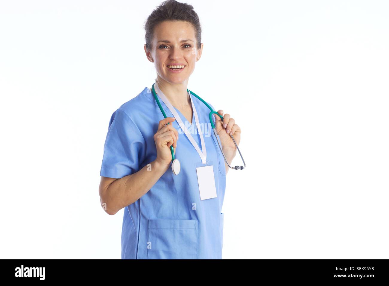 Una donna sorridente medico in scrub blu tiene il suo stetoscopio con entrambe le mani, incarnando esperienza clinica, prontezza e compassionevole Foto Stock