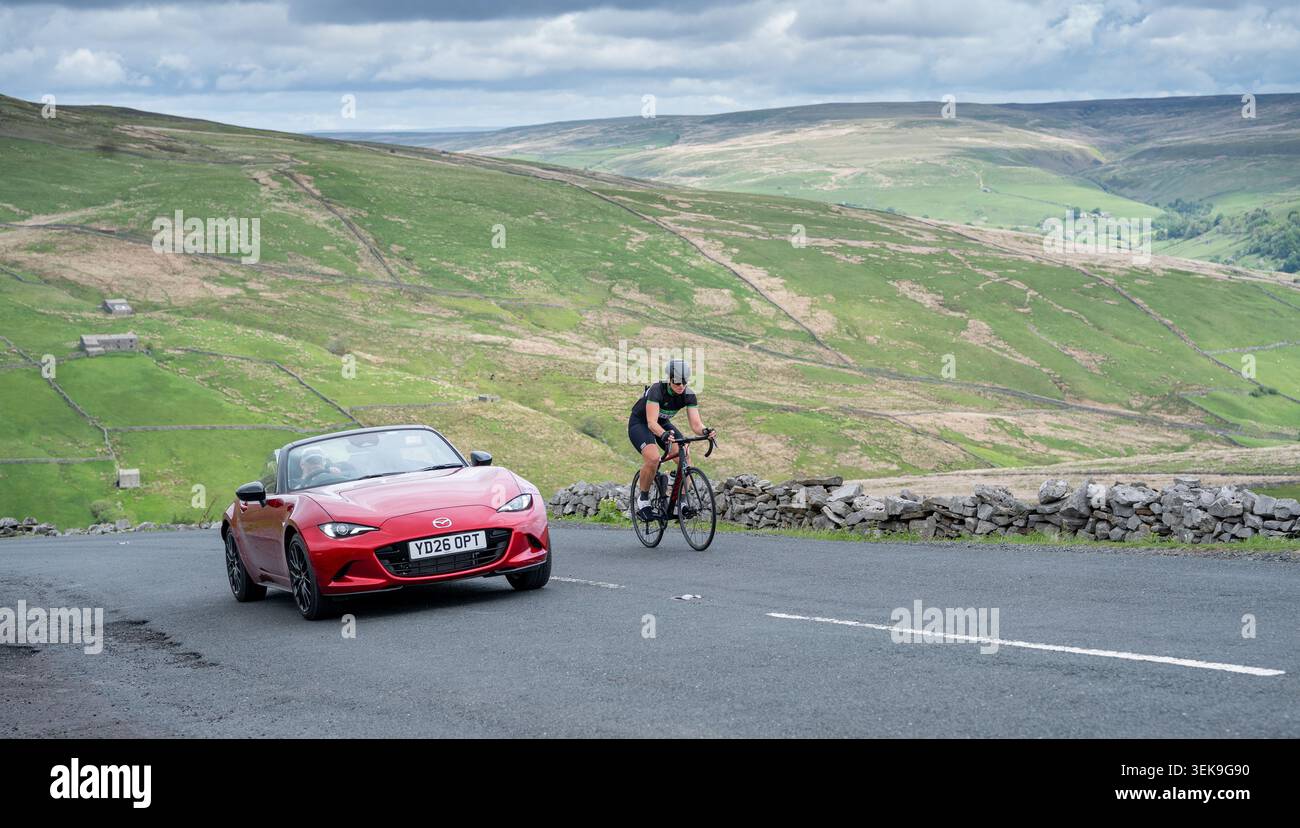 Auto sportiva che sorveglia un ciclista sul passo Buttertubs a Swaledale. Yorkshire Dales National Park, Regno Unito. Foto Stock