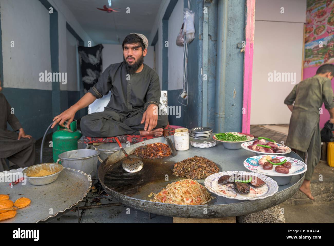 Venditore ambulante afghano che indossa abiti tradizionali preparando vari piatti cucinati su una grande piastra di metallo. Foto Stock