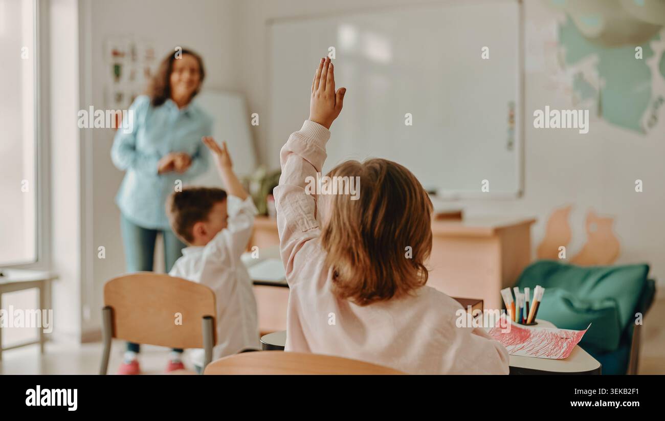 I ragazzi della scuola primaria caucasica alzano la mano durante la lezione con l'insegnante in classe Foto Stock