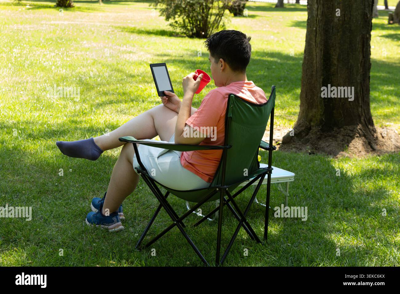 Una giovane mancina beve e legge un e-book in un parco soleggiato, mentre si toglie una scarpa e si flette la gamba Foto Stock