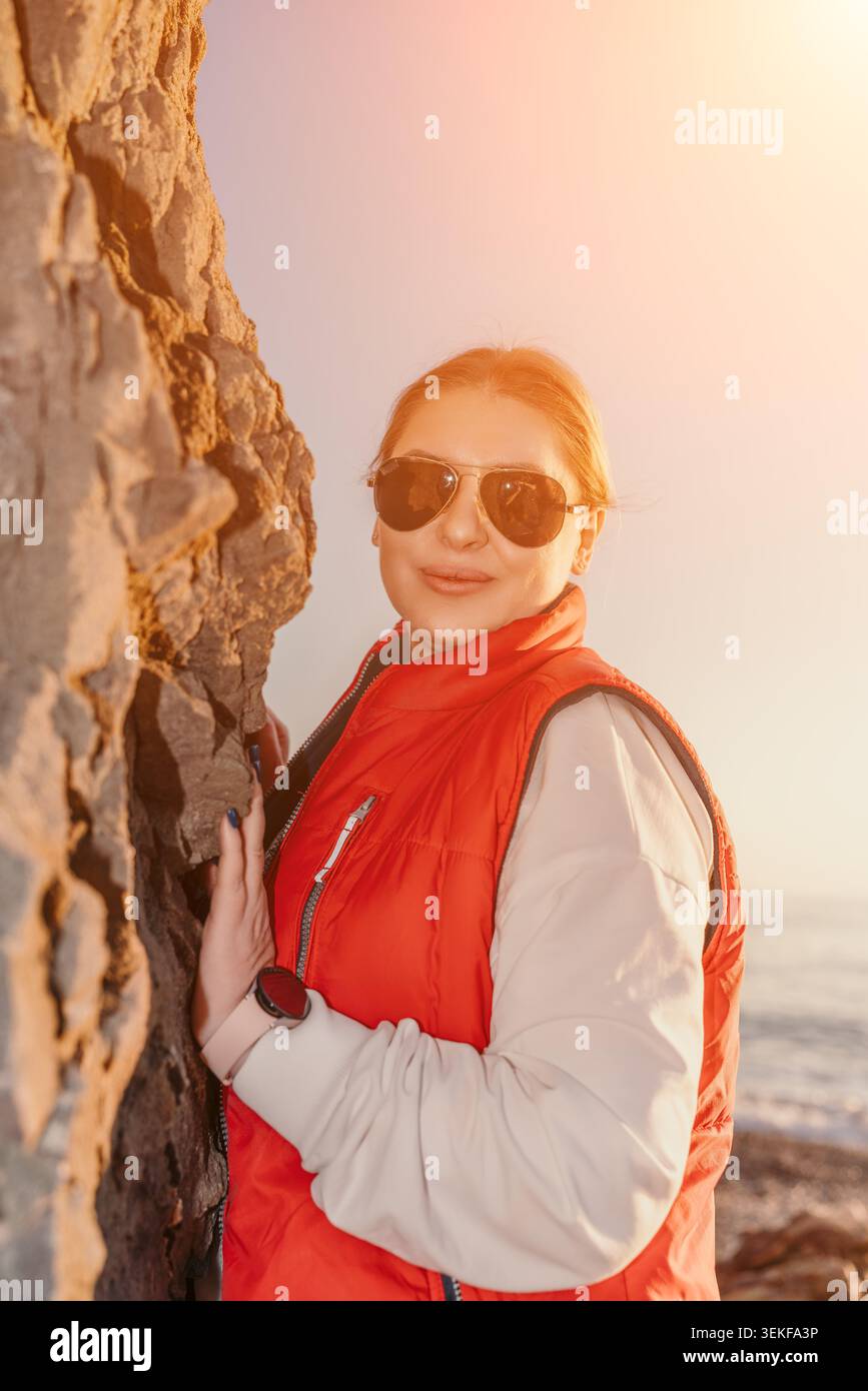 Il viaggio lungo la costa delle donne si staglia davanti a una scogliera rocciosa sulla spiaggia, indossando un gilet rosso e occhiali da sole durante l'ora d'oro Foto Stock