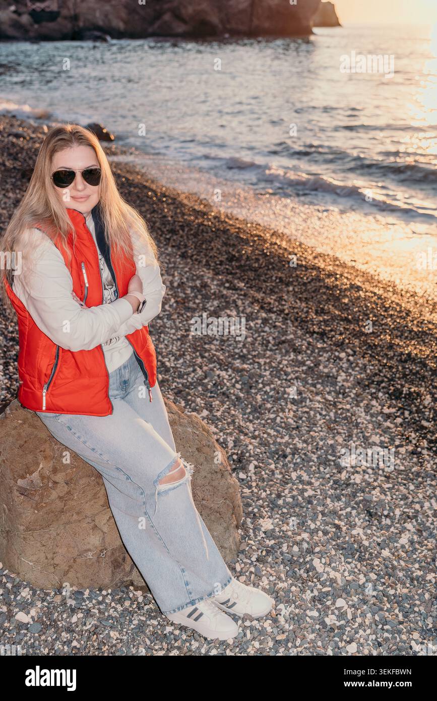 Donna al tramonto sulla spiaggia seduta su una roccia costiera con indosso un gilet rosso e occhiali da sole durante una serata tranquilla in riva al mare Foto Stock