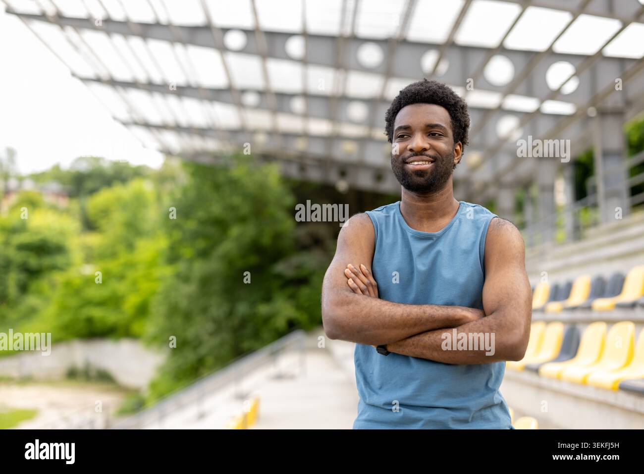 Uomo sicuro di sé sportivo con le braccia incrociate in piedi in uno stadio all'aperto, la sua espressione irradia determinazione e successo, incarna fitness, guarigione Foto Stock