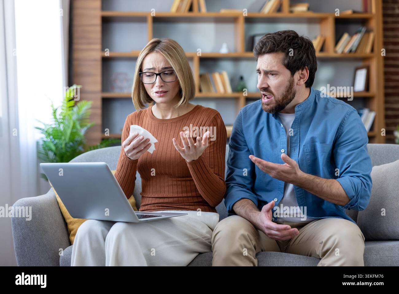 Le difficoltà di relazione emergono mentre una giovane coppia si siede su un divano a casa, con la donna che piange e tiene in mano un fazzoletto mentre guarda un notebook A. Foto Stock