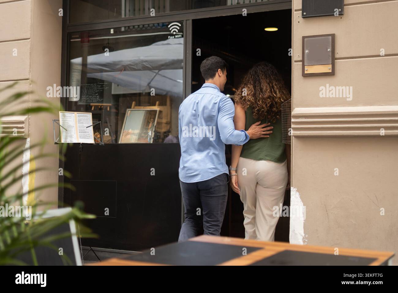 Vista posteriore di una coppia che entra in un ristorante insieme in un ambiente urbano. Uomo che guida delicatamente il suo partner mentre cammina in un locale per un romano Foto Stock