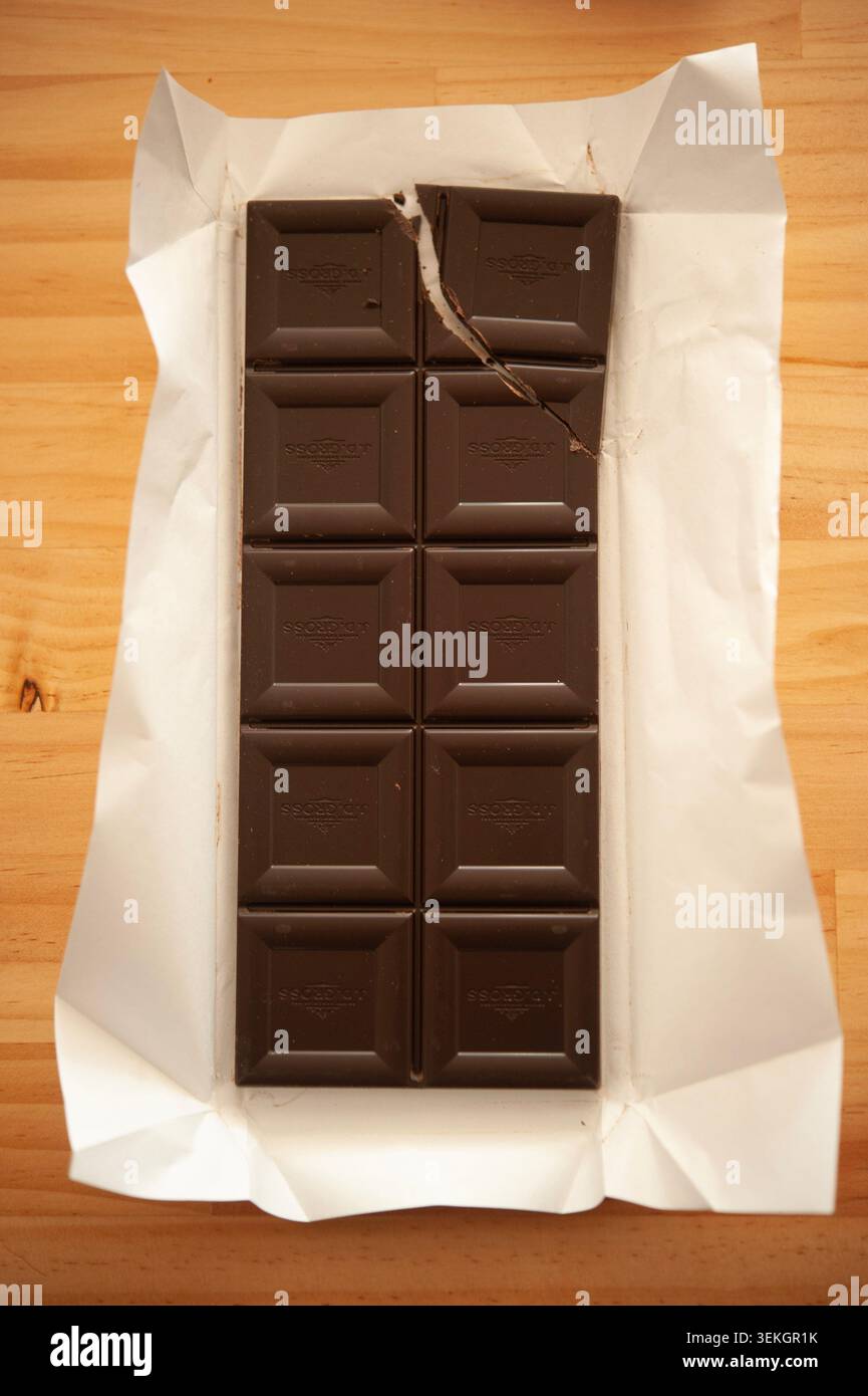 Apri la tavoletta di cioccolato fondente in un involucro bianco su un tavolo di legno, un pezzo rotto, cibo minimalista natura morta Foto Stock