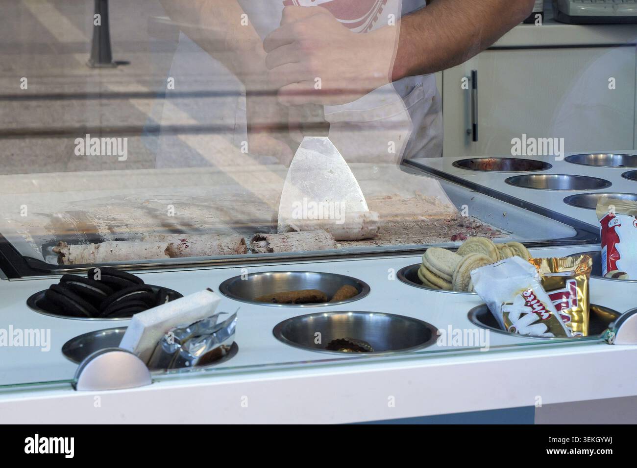 Uomo che raschia e arrotola un gelato cremoso su una padella surgelata, si piega in biscotti e caramelle per creare dessert ghiacciati colorati e ghiacciati in strada Foto Stock
