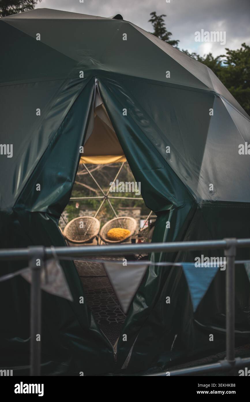 Una tenda a cupola geodetica verde scuro offre uno scorcio nel suo invitante interno, caratterizzato da due sedie intrecciate e un cuscino giallo brillante, accennando a comfor Foto Stock