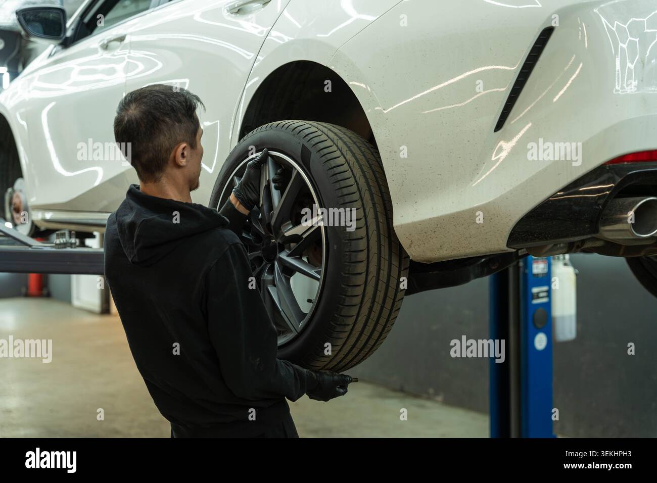 Tecnico che lavora accanto a un'auto bianca su un sollevatore idraulico mentre controlla la ruota posteriore in un'officina di riparazione automobilistica con illuminazione interna brillante Foto Stock