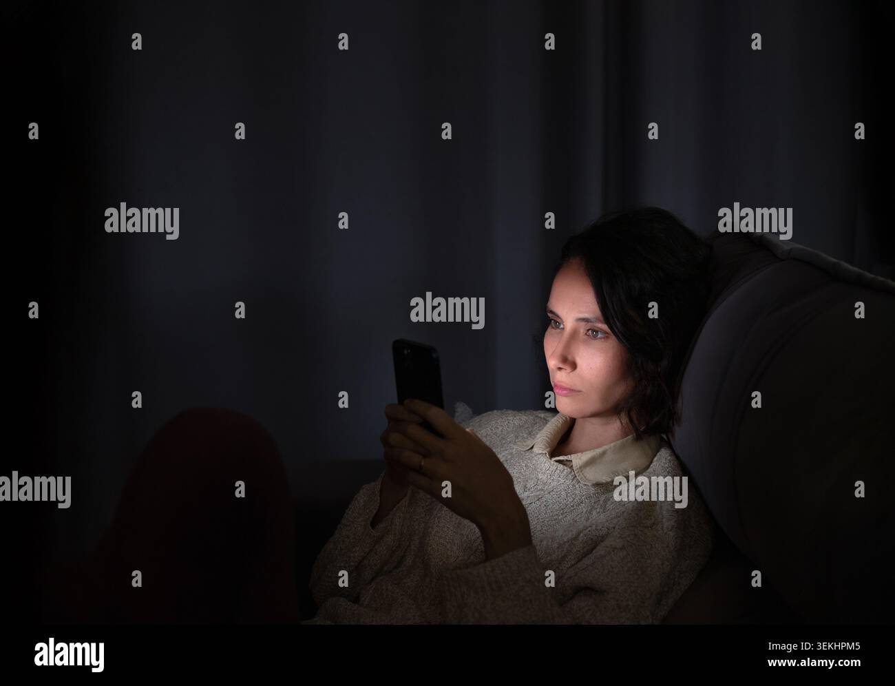 Una donna siede su un divano in una stanza buia, concentrata sul suo smartphone. Sembra ritirata e tesa, catturando sentimenti di ansia e l'impatto di te Foto Stock