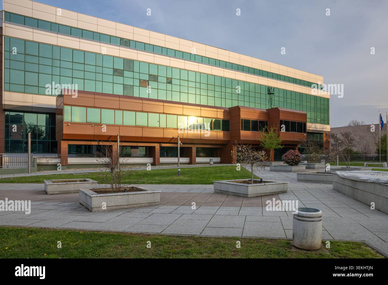 Vinkovci, Croazia - 3 aprile 2026: Moderno edificio di uffici in vetro con facciata verde riflettente che si affaccia su una tranquilla intersezione urbana al tramonto, trasportando Foto Stock