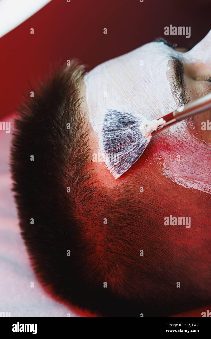 Client receives red light therapy during a cosmetology appointment. A therapist applies a facial product using a brush. The environment has a focused Foto Stock