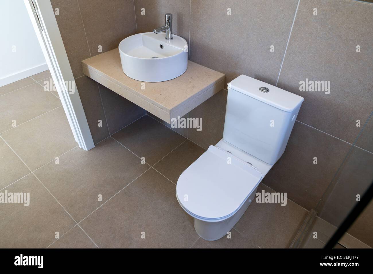 Un bagno pulito e contemporaneo con WC in ceramica bianca e un lavandino rotondo montato su un ripiano galleggiante beige con pareti piastrellate e. Foto Stock