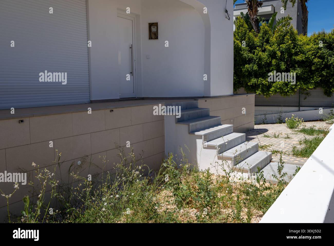 L'esterno di una casa contemporanea presenta una porta bianca, una tapparella e scale di cemento che conducono all'entrata, circondate da verde e pa Foto Stock