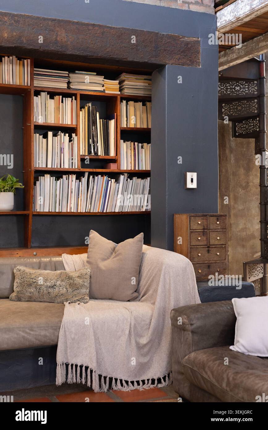 Il divano imbottito è seduto in un salotto in stile loft, con libreria incorporata, tiro e cuscini. Libreria, accogliente, calda, industriale, legno, travi, scale Foto Stock